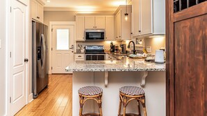 A modern kitchen with white cabinets, stainless steel appliances, a granite countertop, and two wicker stools. - 4005 Rookery 3 Sea Story With 2 Pools Beach Access and Lots of Beach Amenities (Gulf Shores)