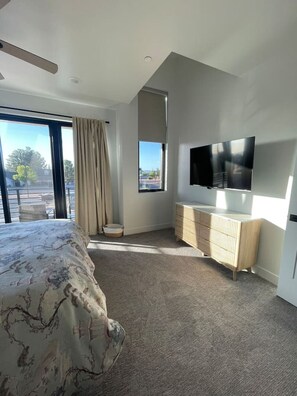 A modern bedroom with a large bed, a TV mounted on the wall, and a view of the outdoors through a window.