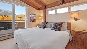 A bedroom with a large bed, wooden ceiling, two bedside lamps, a nightstand, and a view of autumn trees and mountains.