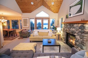A spacious living room with a stone fireplace, a large window overlooking a scenic view, a cozy sofa, and a dining area. - Charming 3-bedroom house in Vashon with cool AC breeze (Vashon)