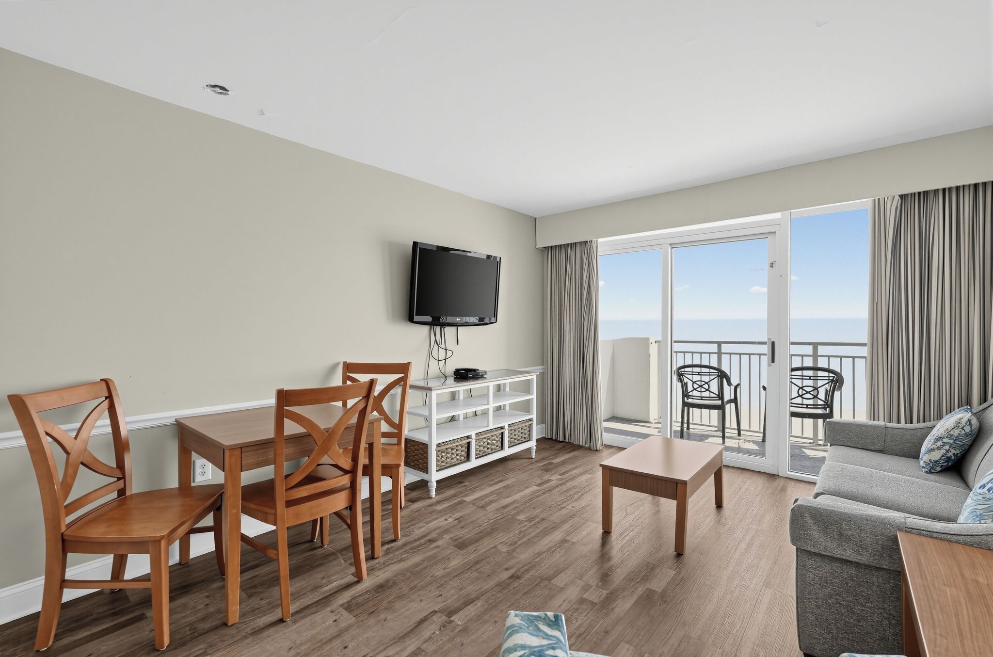 A living room with a TV, wooden furniture, a balcony with chairs, and a view of the sea.