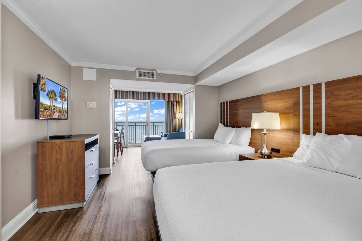 Ein Hotelzimmer mit zwei Betten, einem an der Wand montierten Fernseher, einem Schreibtisch mit Stuhl und einem Balkon mit Blick zum Himmel.