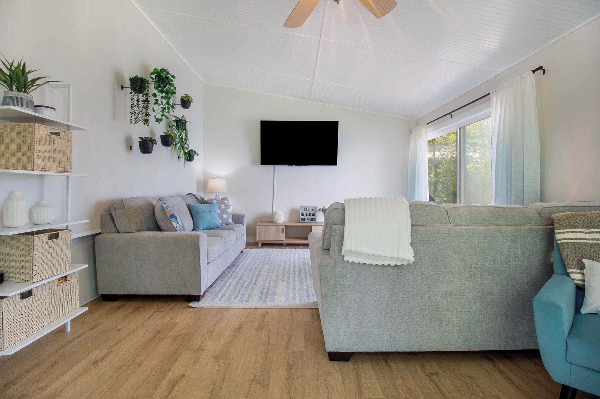 Een moderne woonkamer met een grijze bank, een blauwe fauteuil en een flatscreen televisie aan de muur. Er staan rieten manden op planken en diverse potplanten op een wandplank.