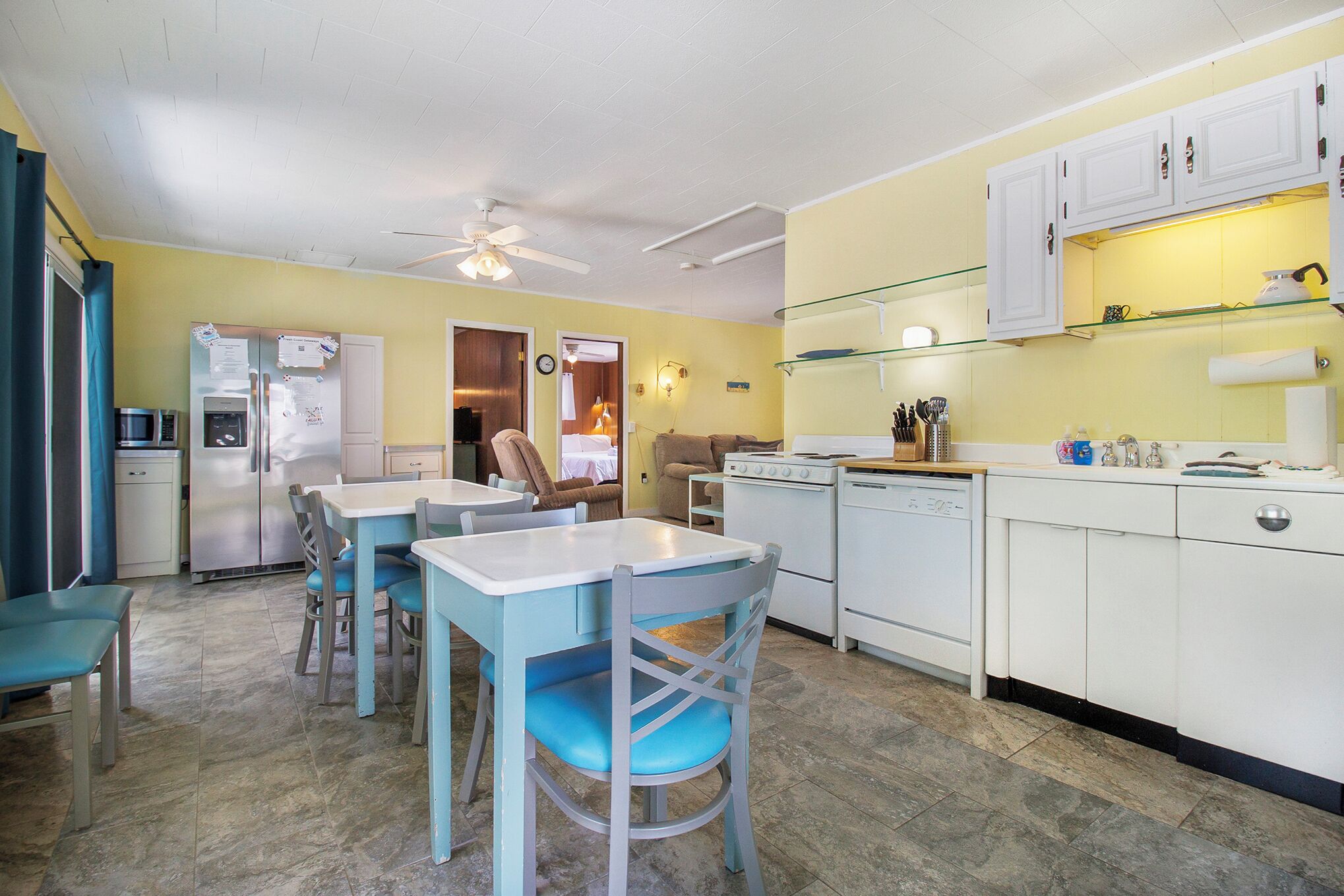 A compact kitchen with white cabinets, a dining area with blue chairs, and a ceiling fan.
