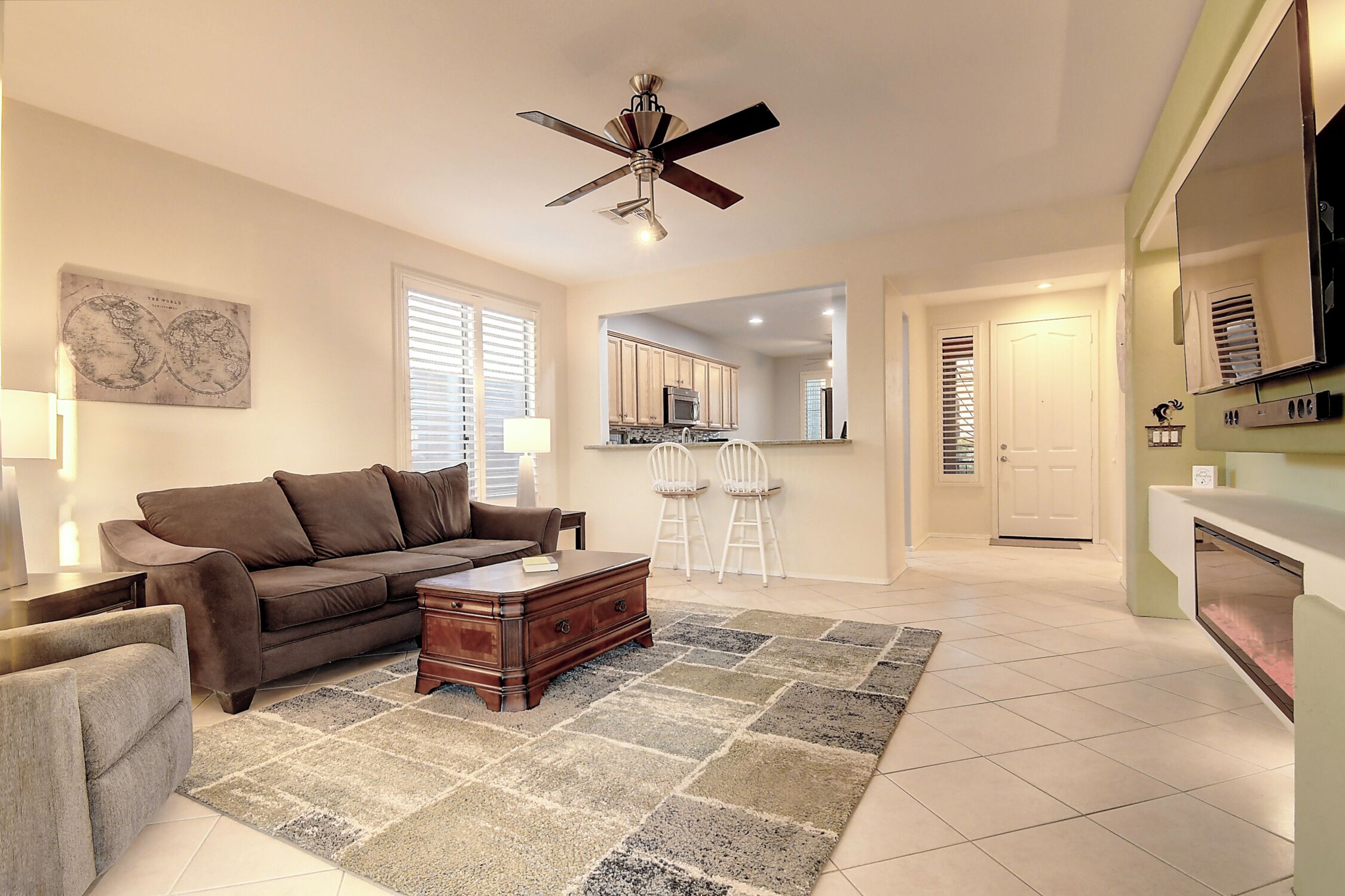 A living room with a brown sofa, a wooden coffee table, and a patterned rug. There is a ceiling fan, a kitchen area with a microwave, and a large mirror.