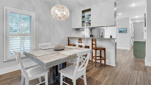 A modern kitchen with white cabinetry, a wooden dining table with white chairs, and a hanging light fixture. - The Grey Gull (Panama City Beach)