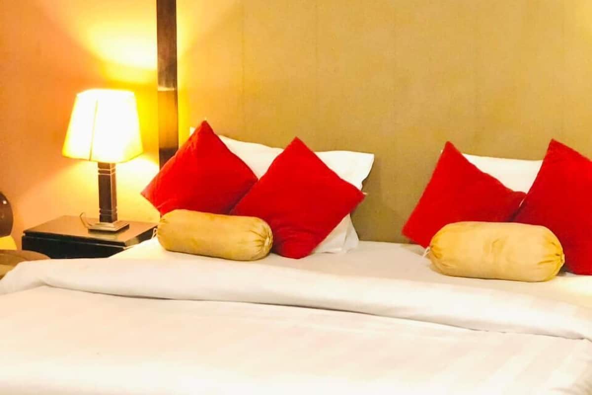 A hotel room with a large bed, red and beige pillows, a bedside lamp, and a nightstand.