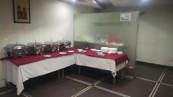 Un buffet avec des plats et des récipients couverts disposés sur une table, un panneau « Interdiction de fumer » sur une cloison en verre et un sol carrelé.
