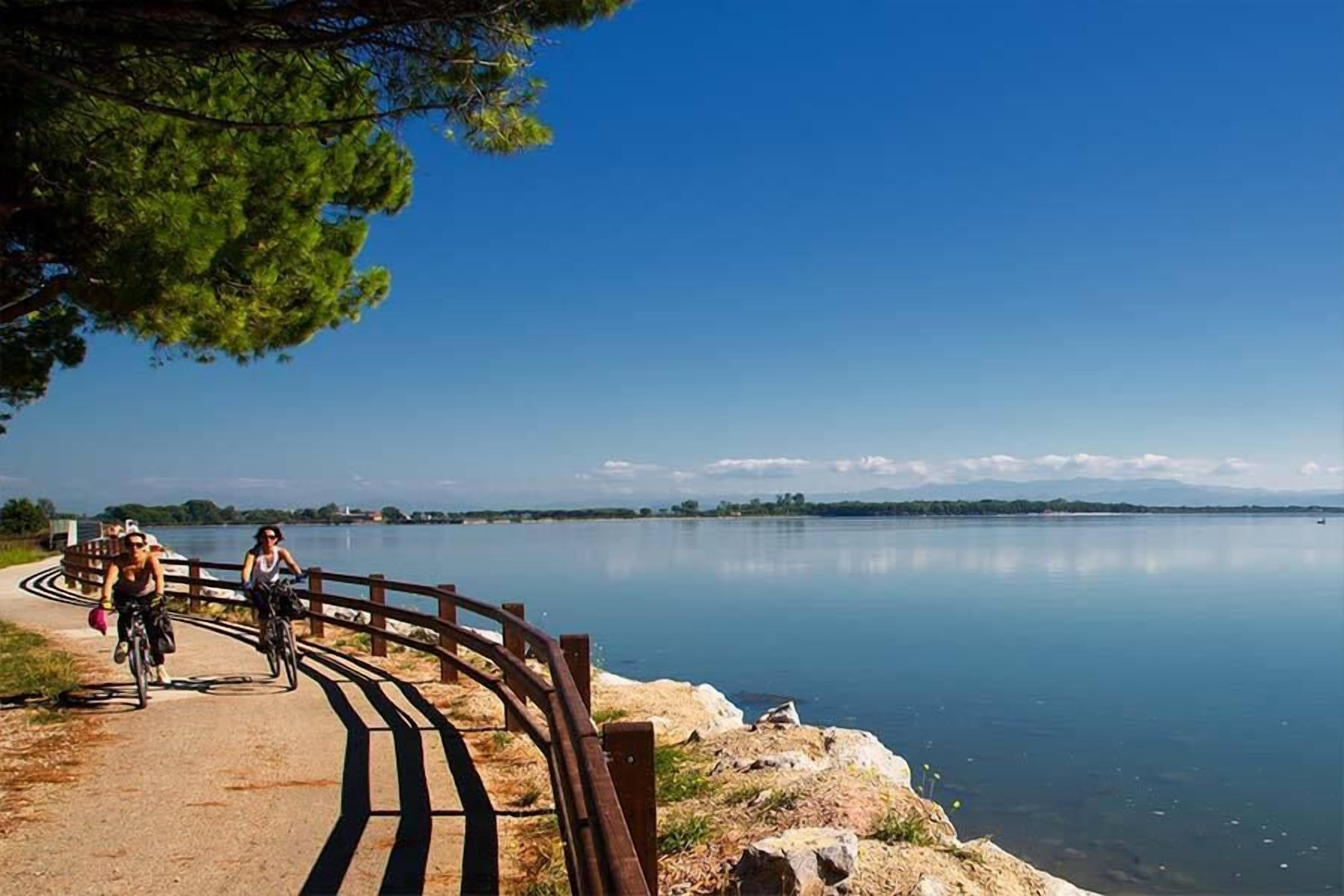 Un sentier pavé longe un lac, bordé d’une rambarde en bois. Le ciel est dégagé et l’on aperçoit des montagnes au loin.