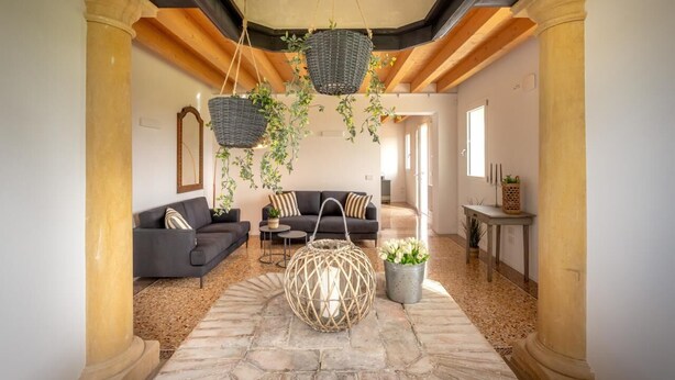 Amplia sala de estar con piso de piedra, sofá de color oscuro, una mesita de centro redonda de fibras naturales y cestas colgantes con plantas.