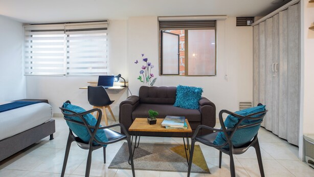 A compact living space with a bed, a sofa, a coffee table, two chairs, a desk with a computer, and a wardrobe.