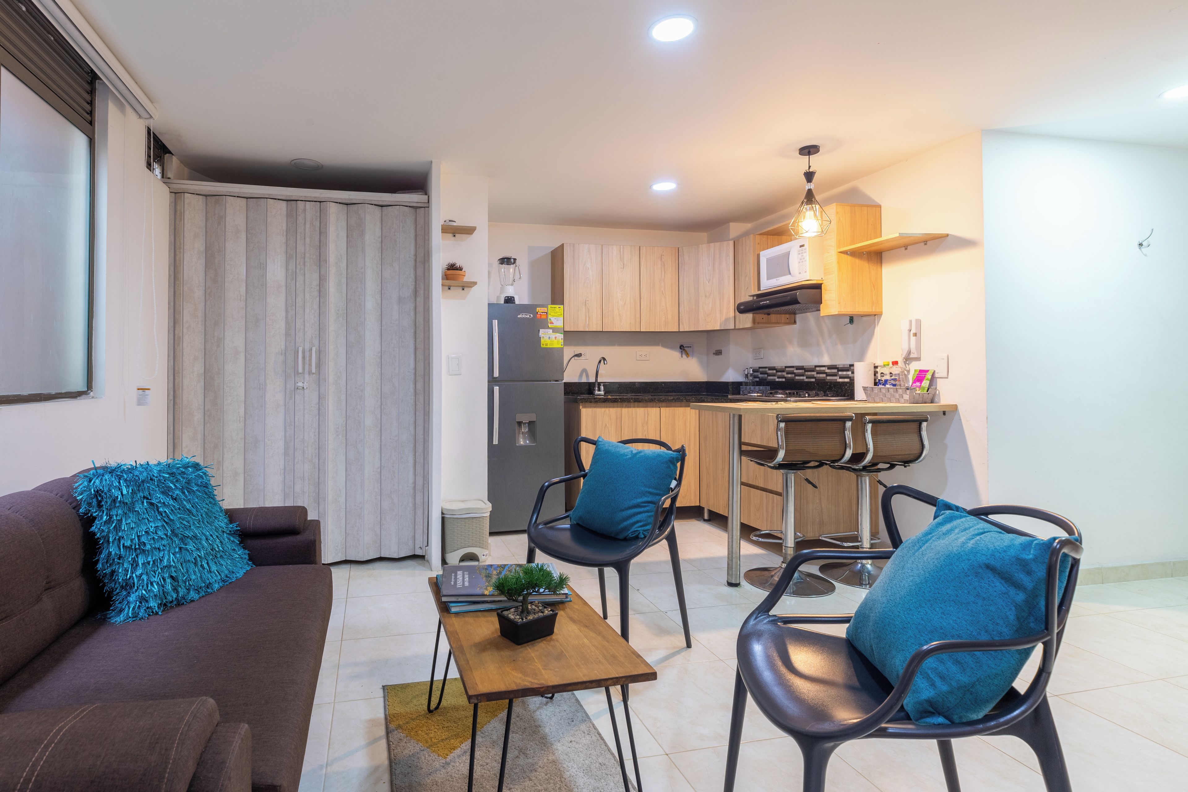 A modern kitchen with wooden cabinets, a refrigerator, and a microwave. There is a dining area with two chairs and a table, and a living room with a sofa and a coffee table.