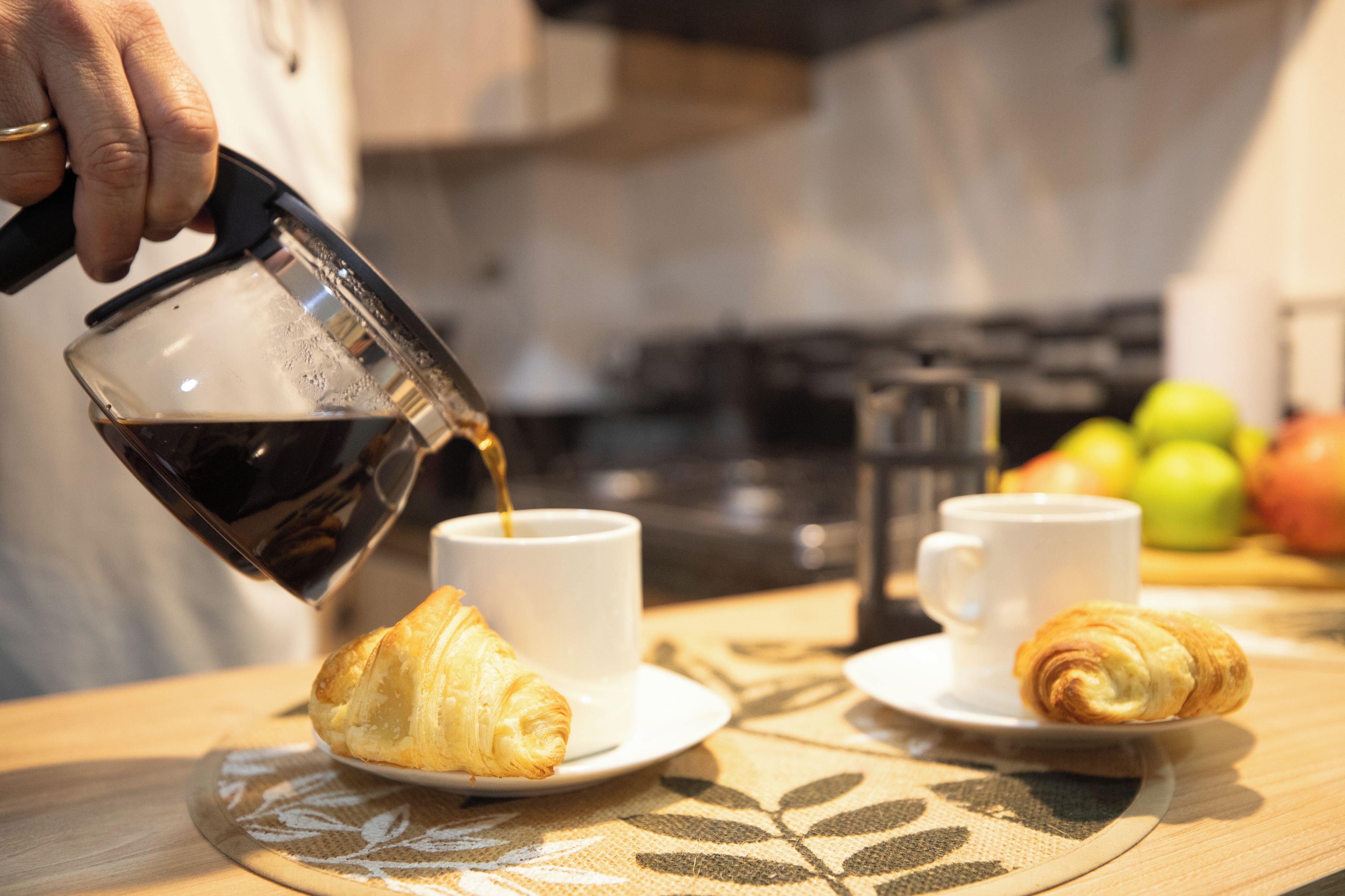Una persona sirviendo una bebida en una taza, con croissants y manzanas sobre la mesa.