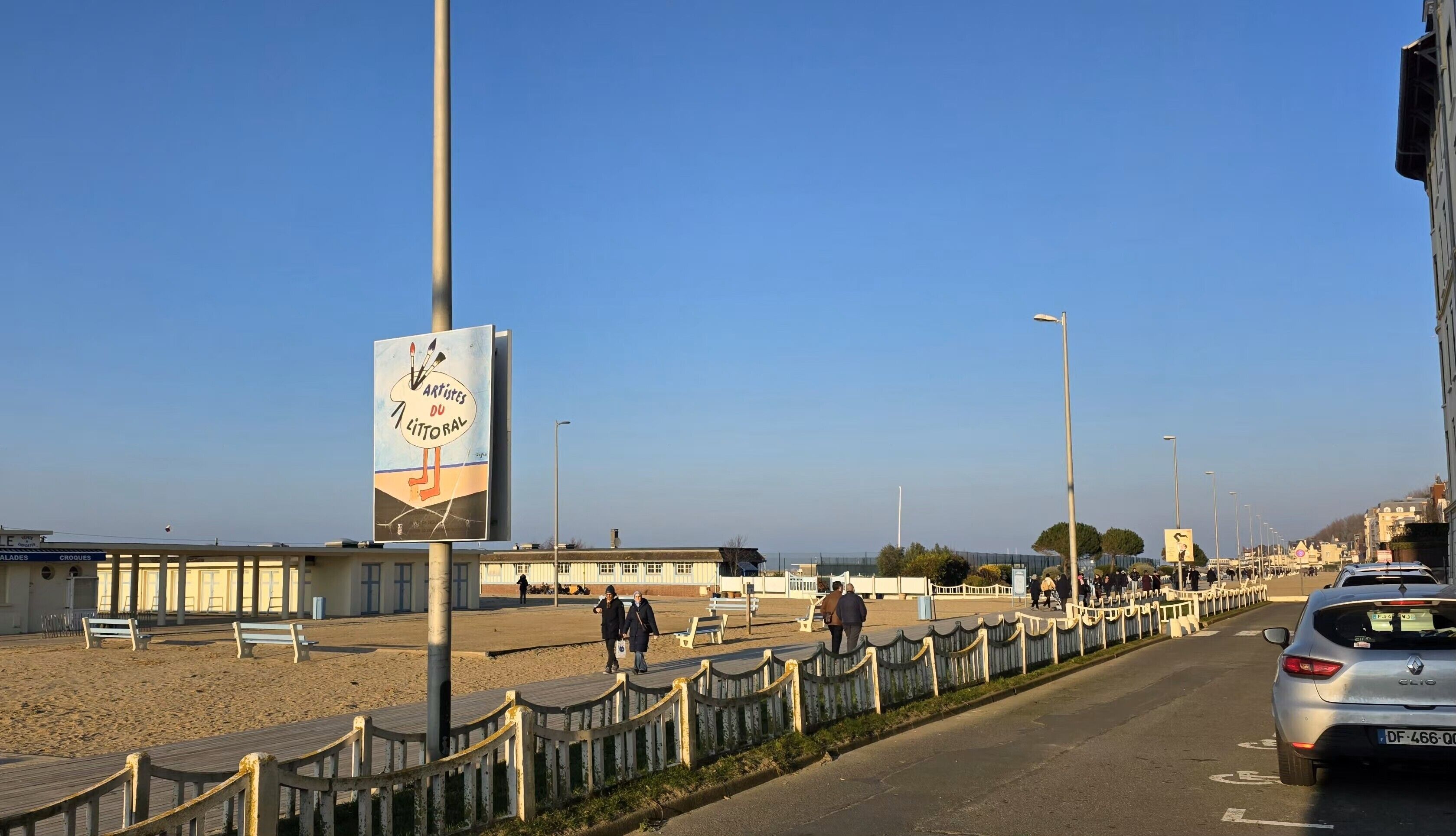 Une promenade en bord de mer agrémentée d’une jetée en bois, de bancs et d’un panneau publicitaire annonçant « Artistes du Littoral ».