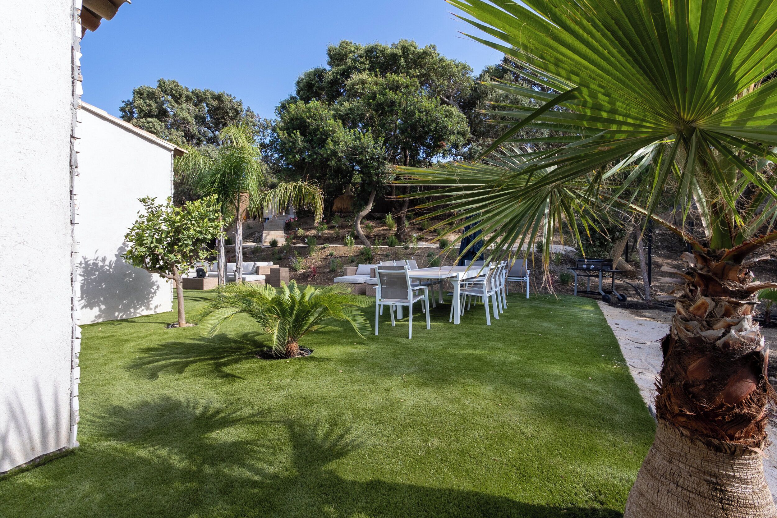 Une terrasse extérieure bien entretenue, avec une table et des chaises, entourée d’une végétation luxuriante et bordée d’un bâtiment blanc.