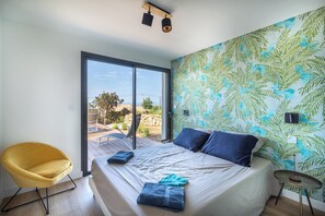 A bedroom with a bed, a yellow chair, a small table, and a view of the outdoors through a glass door. - Villa de La Madrague – Presqu’île de Giens (Hyères)