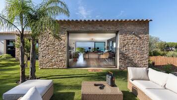 A modern house with a stone exterior, a glass door leading to a living area with a view, and outdoor furniture.