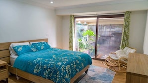 A bedroom with a bed, a wooden nightstand, a wicker chair, a wooden table, and a sliding glass door leading to a balcony with plants.