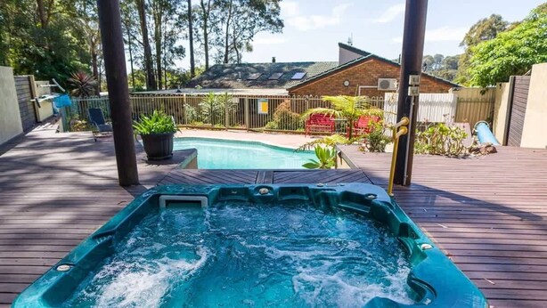 A hot tub on a wooden deck with a pool in the background, surrounded by a fenced yard with plants and a house.