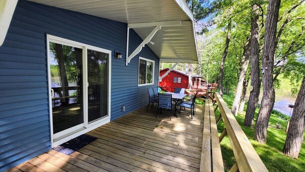 A blue house with a deck, a red cabin, and a wooden fence. - Arcade Room + Fire Pit + Creek Views | Near Waterparks (Wisconsin Dells)