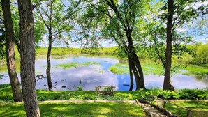 A tranquil pond surrounded by trees, a wooden bench, and a wooden fence. - Arcade Room + Fire Pit + Creek Views | Near Waterparks (Wisconsin Dells)