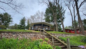 A modern building with a wooden staircase leading up to a deck, surrounded by greenery and trees. - Arcade Room + Fire Pit + Creek Views | Near Waterparks (Wisconsin Dells)