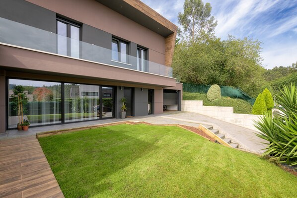 A modern two-story house with a large glass balcony, a well-maintained lawn, and a stone pathway leading to the garage.