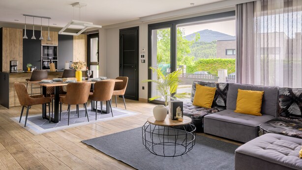 A modern living room with a dining area, a sofa with yellow cushions, and a view of greenery and mountains through large windows. - Detached House with Landscape Views and Charm in the Heart of the Basque Country (MARKAIDA)