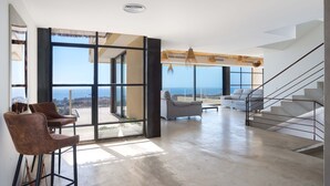 Amplia sala de estar con un ventanal que da al océano, una escalera y un área de descanso con sofá y sillones.