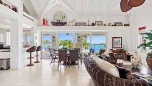 A spacious living area with a large window overlooking the sea, a dining area with wicker furniture, and a kitchen visible in the background. - Villa 3: islandpropertiesonline: 5-bedroom villa infinity pool & terrace (Maho)