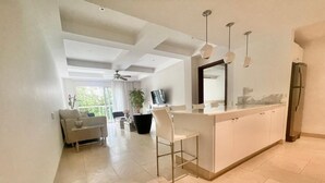 Et moderne køkken med en central ø, et spiseområde med en sofa og et stort vindue, der giver naturligt lys.