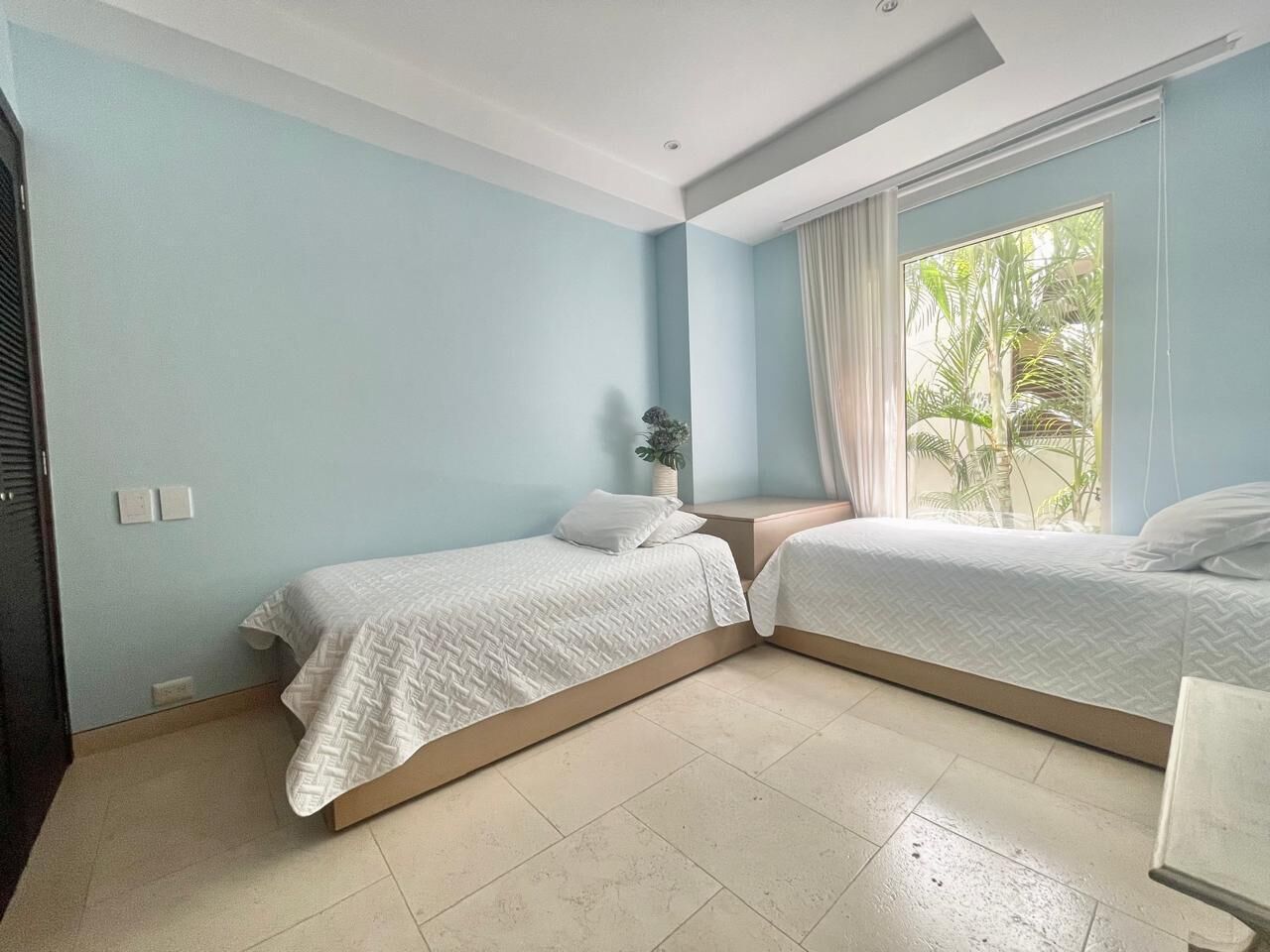 A bedroom with two beds, light blue walls, a window with curtains, and a small plant on the nightstand.