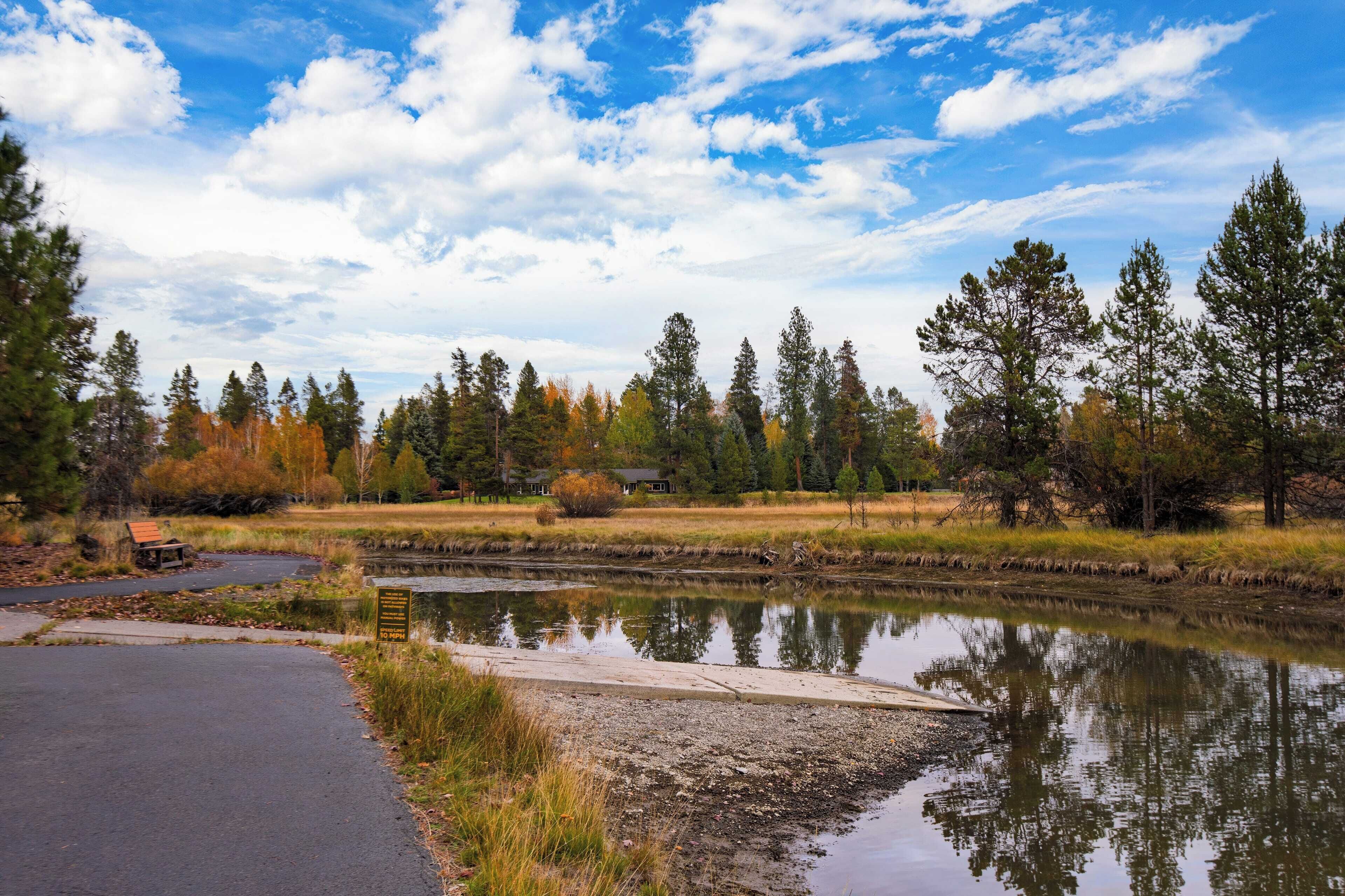 A serene lakeside view with a paved path, a wooden bench, and a small house in the background.