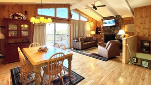 A spacious living room with a wooden dining table, chairs, and a fireplace. There is a television mounted on the wall and a ceiling fan. - Beautiful Cabin in Chalet Village. Hot Tub, 3 Decks, Stunning Mountain Views  (Gatlinburg)