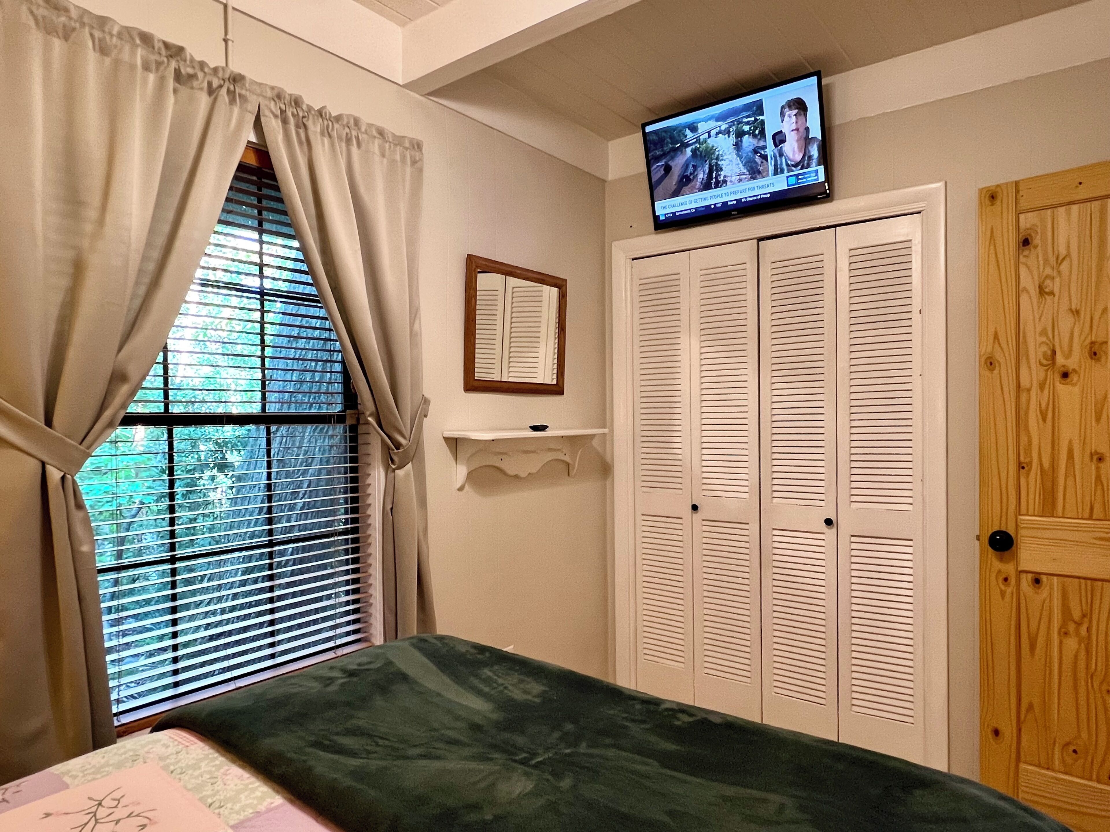 Une chambre à coucher avec un lit, un téléviseur fixé au mur, une fenêtre avec des rideaux et une porte en bois.