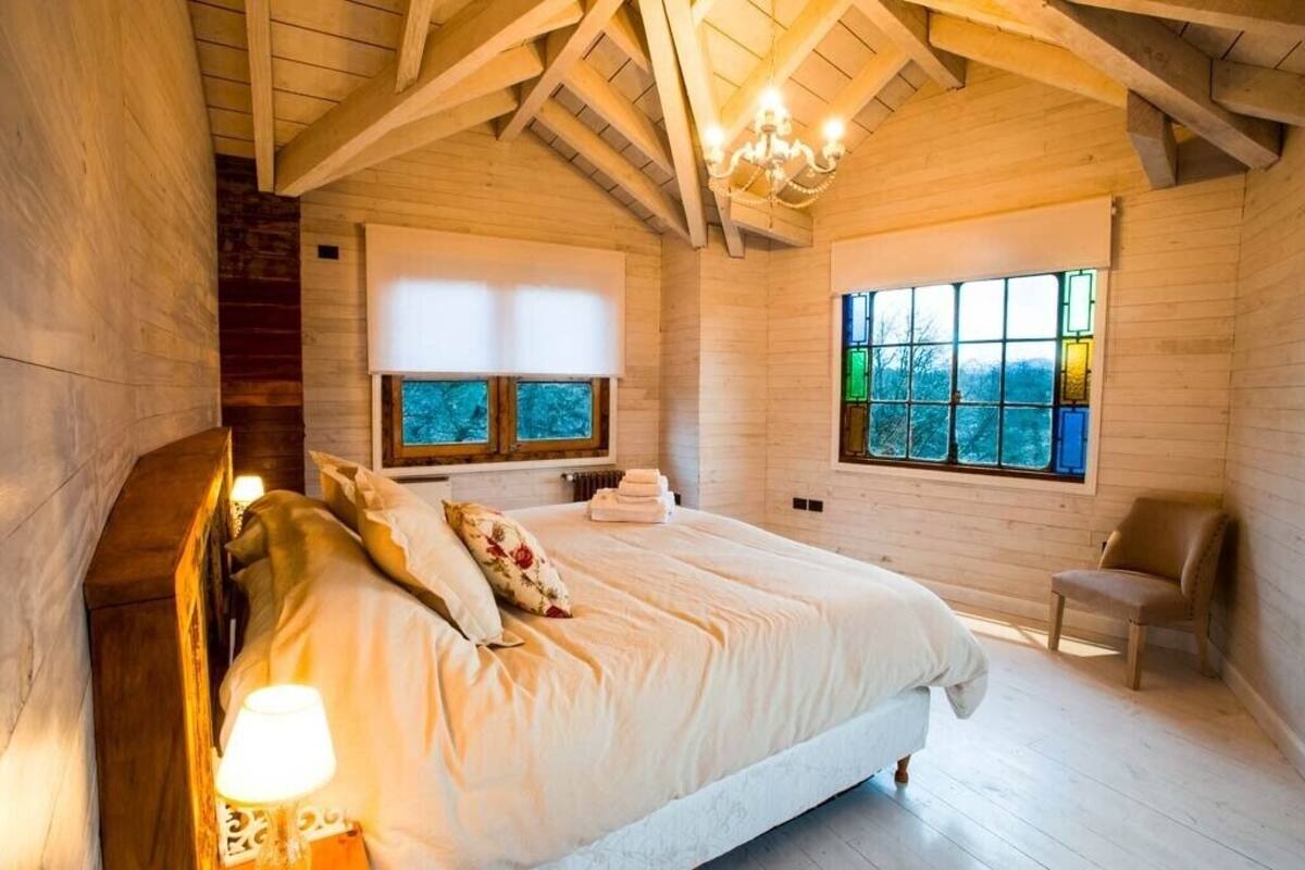 A cozy bedroom with a wooden ceiling, a large bed with pillows, a nightstand, and a window with a view.