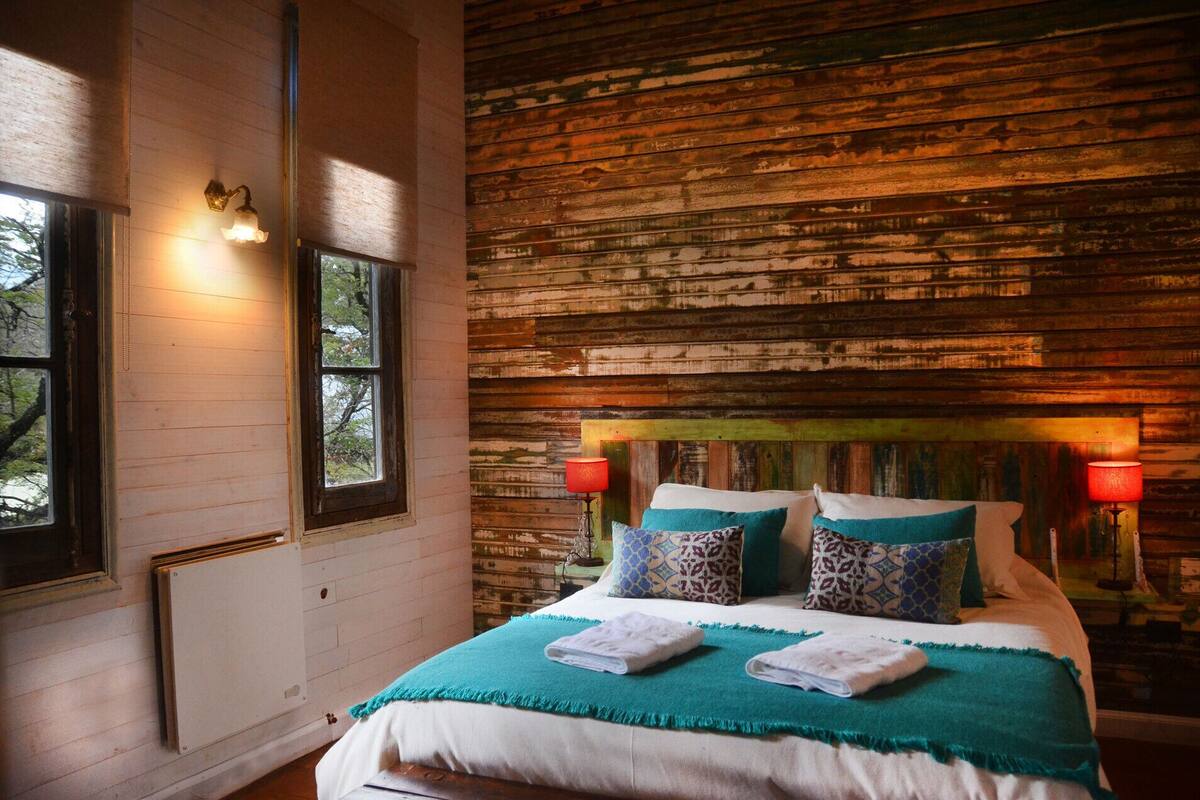A rustic bedroom with a wooden headboard, a bed with multiple pillows, two bedside lamps, and a window with a view of trees.
