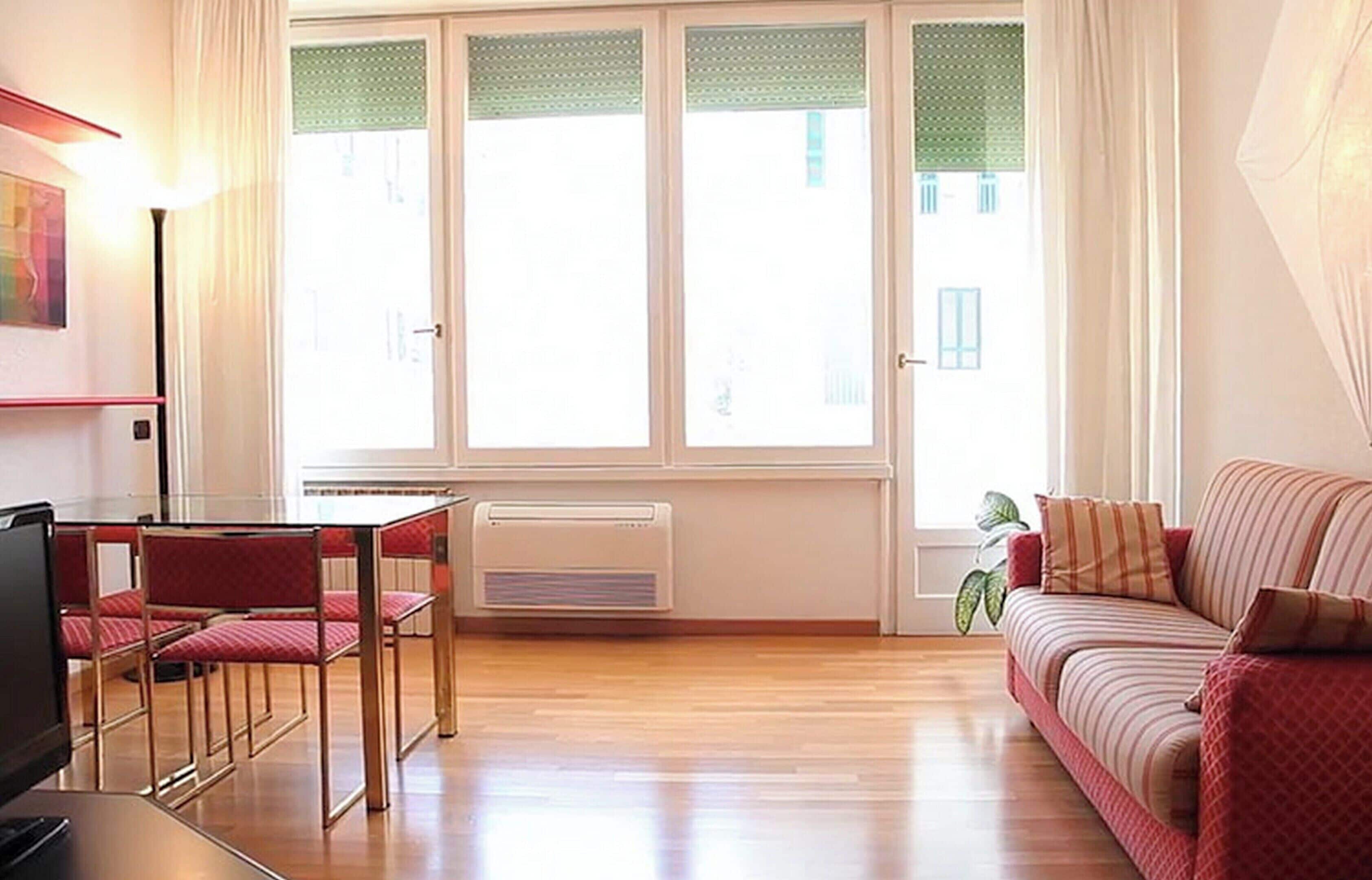 A room with a wooden floor, a red sofa, a dining table with chairs, and a large window with curtains.