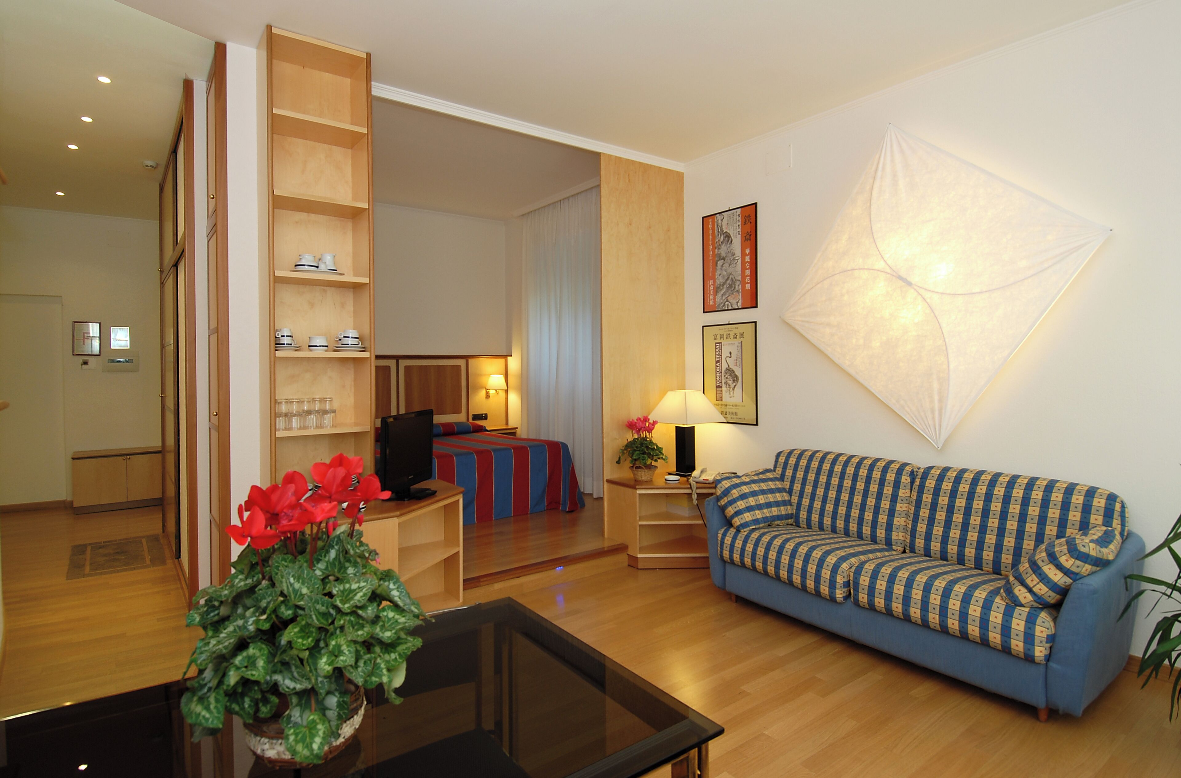 A hotel room with a blue sofa, a glass coffee table, a wooden sideboard, a bed with a striped bedspread, a wall-mounted lamp, and a framed picture.