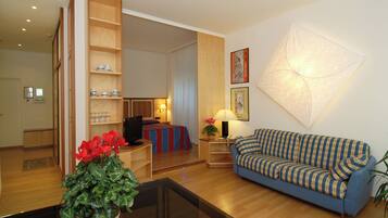 A hotel room with a blue sofa, a glass coffee table, a wooden sideboard, a bed with a striped bedspread, a wall-mounted lamp, and a framed picture.