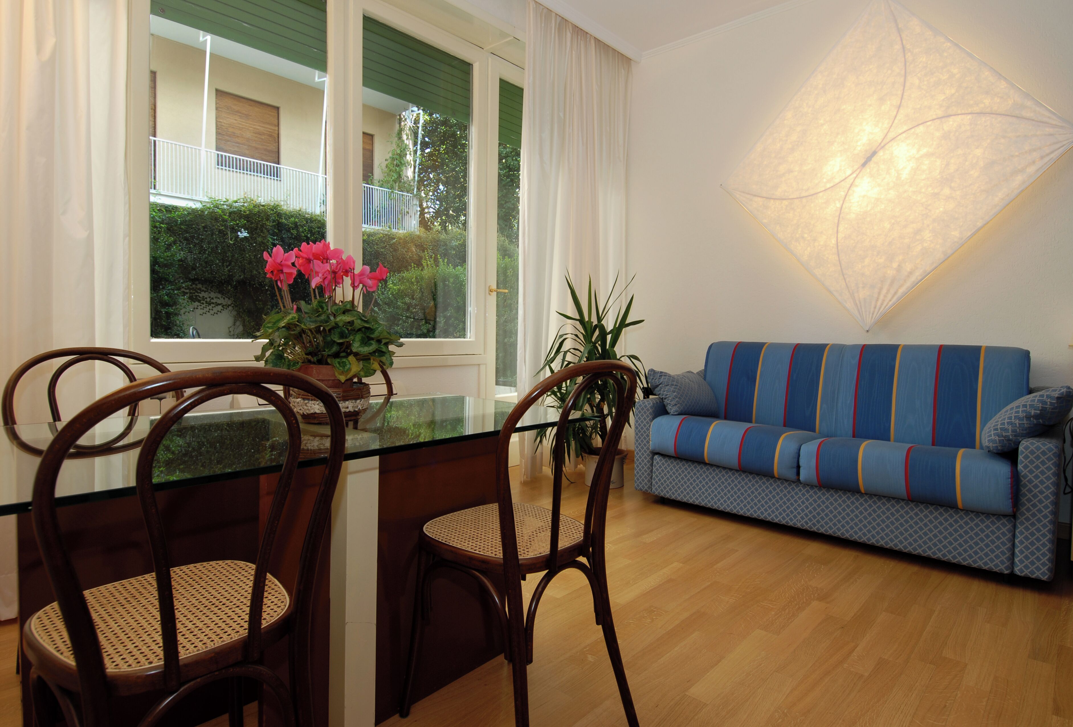 A modern living room with a glass dining table, two wooden chairs, a blue sofa, and a large window with curtains.