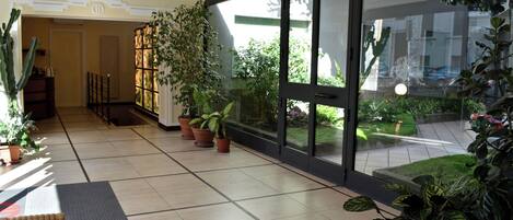 A hotel lobby with a tiled floor, potted plants, a wicker chair, and a glass door leading to an outdoor garden.