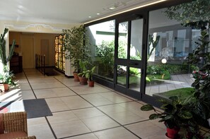 A hotel lobby with a tiled floor, potted plants, a wicker chair, and a glass door leading to an outdoor garden. - Prati Home (Rome)