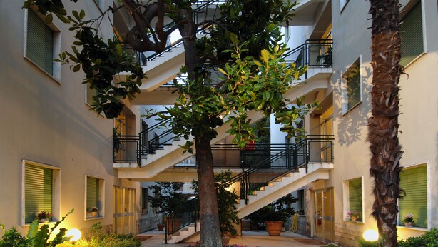 Un edificio residencial de varios niveles con un patio central que presenta un árbol, plantas en macetas y un camino empedrado.