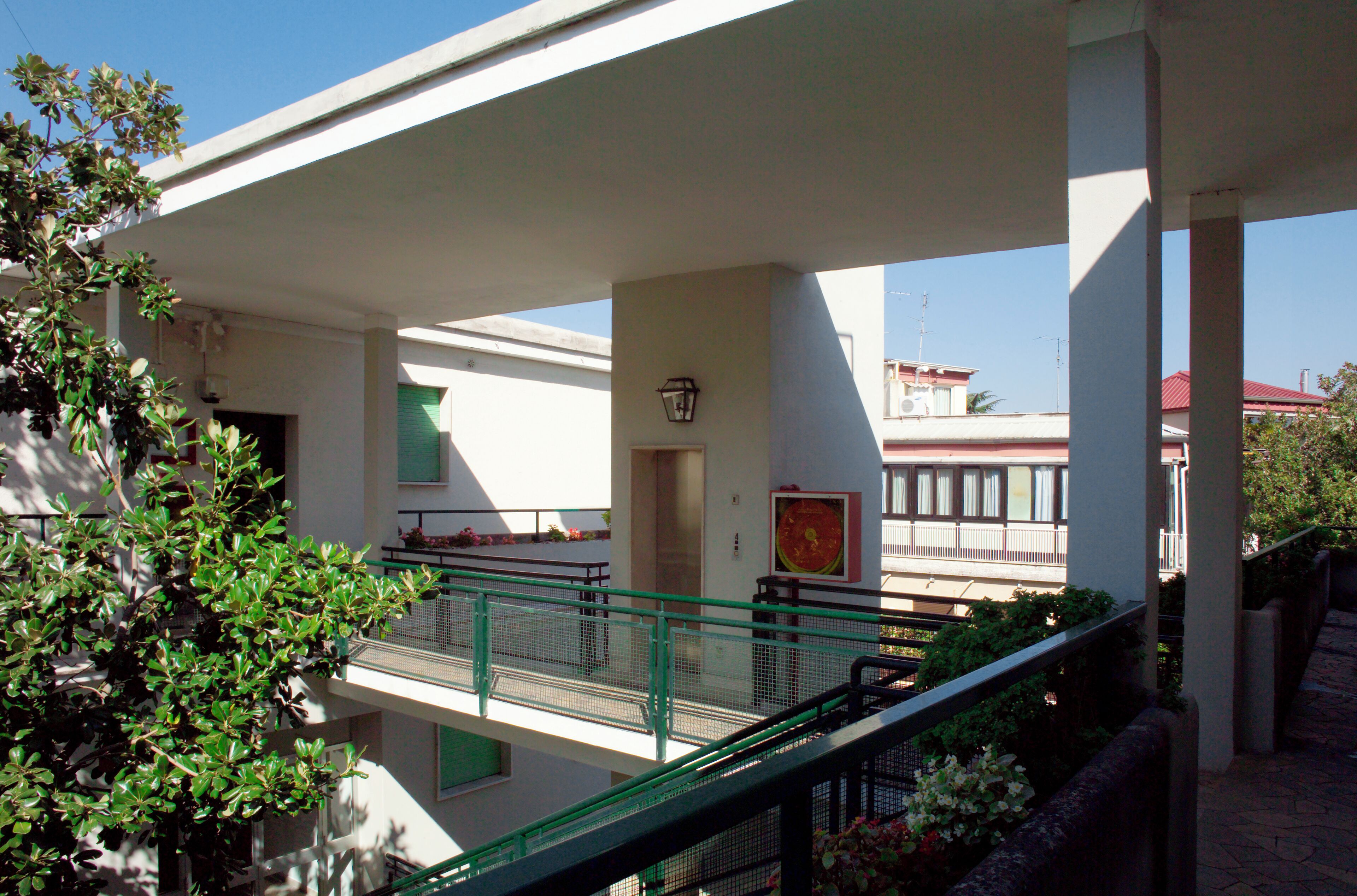 A covered outdoor area with a green metal railing, a white building with a red roof, and a tree with green leaves.
