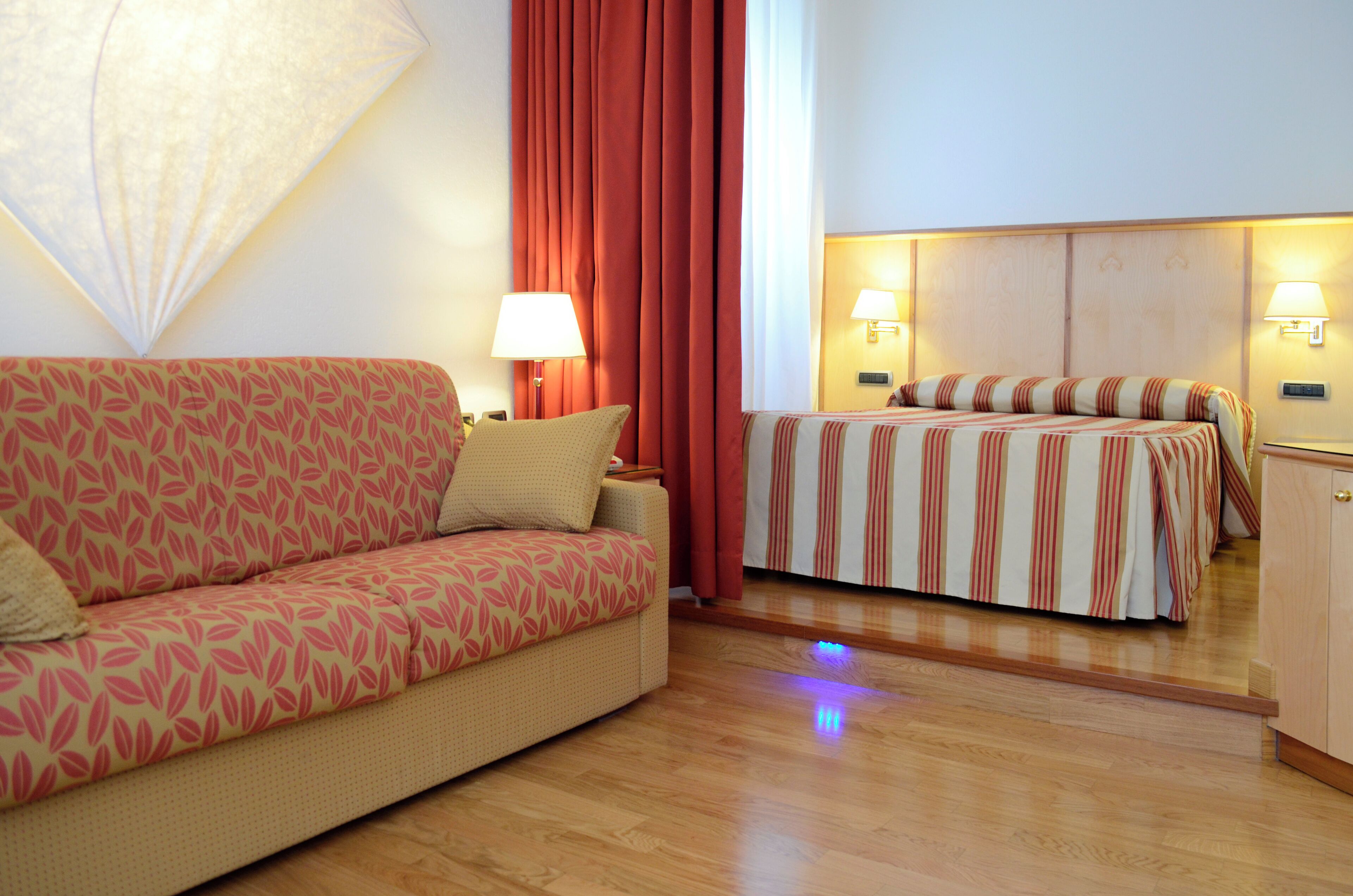 A hotel room with a sofa, a bed with striped bedding, two bedside lamps, and a wooden floor.