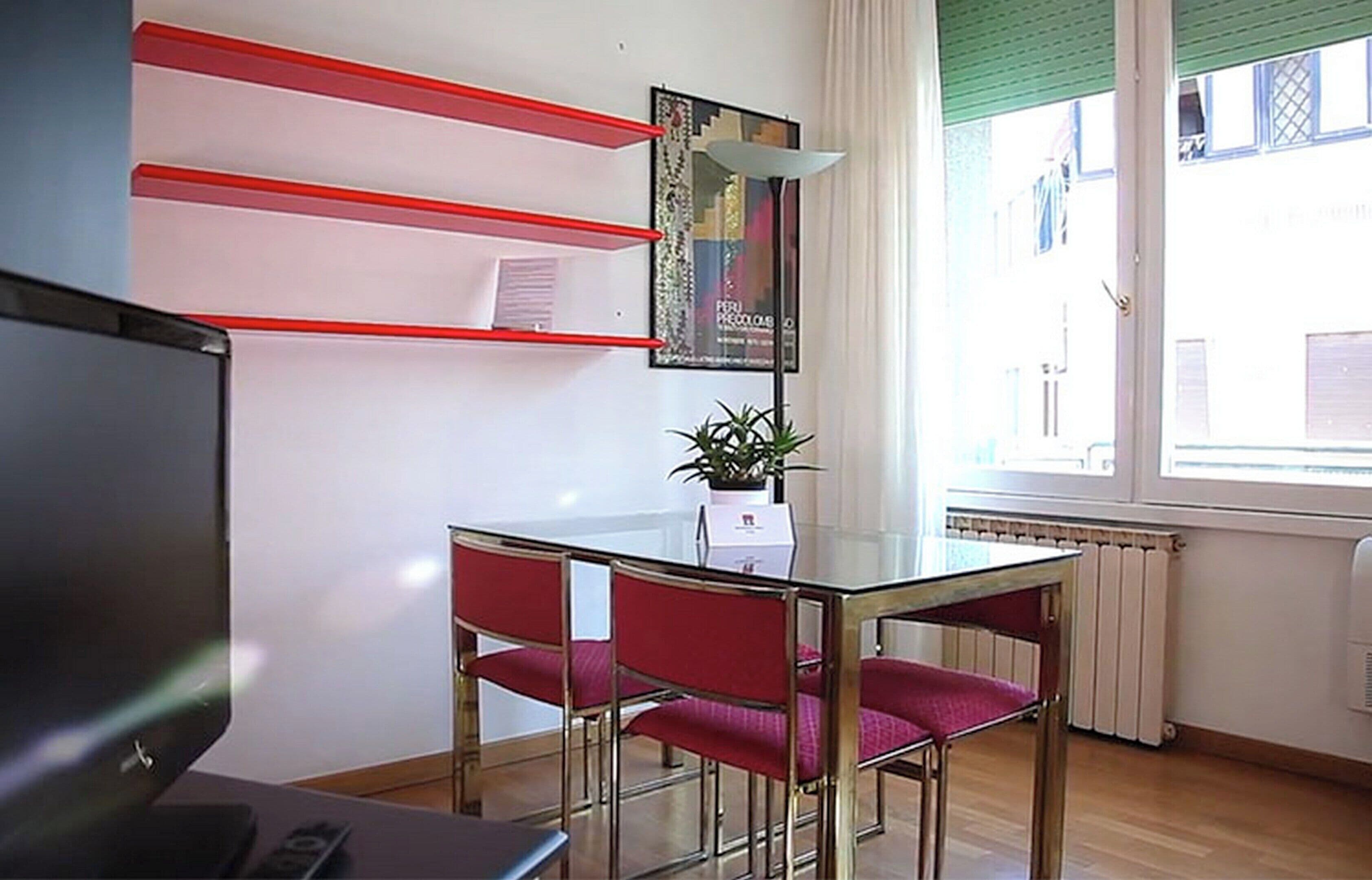 A room with a glass table, red chairs, a potted plant, a poster on the wall, and a window with blinds.