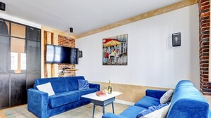 Una sala de estar con un sofá azul, un televisor de pantalla plana montado en una pared de ladrillo y un cuadro de una escena callejera.
