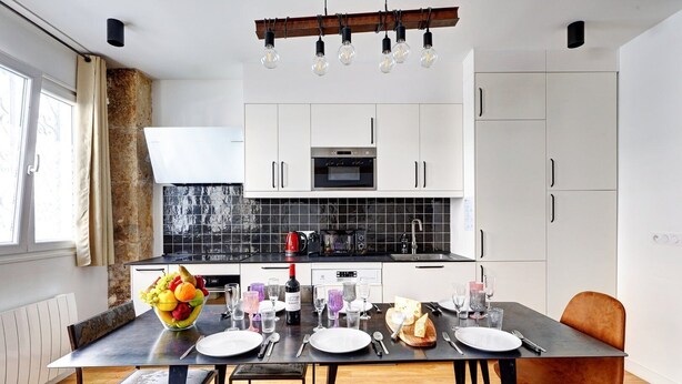Una cocina moderna con armarios blancos, salpicadero negro y una zona de comedor con una mesa dispuesta para cuatro comensales.