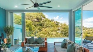 Ein modernes Zimmer mit einem großen Glasfenster zum Pool, einem Deckenventilator und Blick auf eine üppige Landschaft.